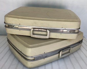 May include: Two vintage off-white hard-shell suitcases with silver accents and the word "Hartmann" on the top suitcase.