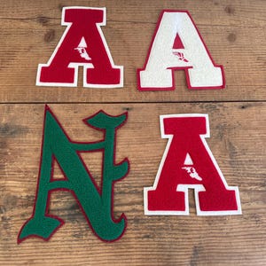 May include: Four felt letter patches arranged in a group. Three patches are the letter A in a block font, with two in red and one in white. The fourth patch is the letter N in a green, stylized cursive font with a red border outline.