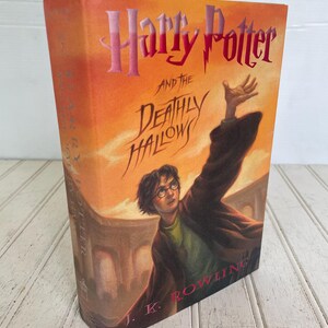 May include: A hardcover book titled "Harry Potter and the Deathly Hallows" by J.K. Rowling. The cover features an illustration of Harry Potter with his arm raised against an orange and yellow sky. The title is in pink and the author's name is in red.