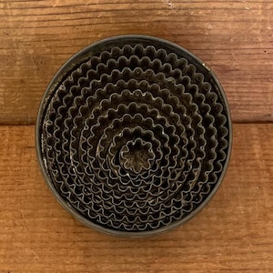 May include: A set of vintage, nested, fluted metal cookie cutters. The cutters are arranged in a circular pattern, with the smallest cutter in the center. The metal has a dark, aged appearance, suggesting it is antique. The cutters are on a wooden surface.