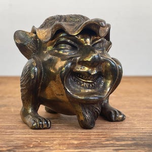 May include: A bronze-toned sculpture of a laughing, mythical creature with a large open mouth and detailed facial features. The sculpture has a textured surface and is posed on all fours, with clawed feet. The creature's head is adorned with a ruffled headdress.