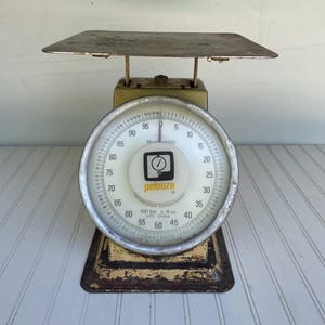 May include: An antique Pelouze Model 40100 kitchen scale with a rectangular metal platform and a circular dial. The dial displays measurements up to 100 lbs and 4 oz. The scale has a worn, vintage appearance with a yellow and brown color scheme.