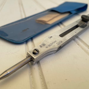 May include: A silver metal compass with a blue leather case. The compass has a sliding mechanism to adjust the length of the compass. The compass is labeled "Alvin W. Germany Line Circle".