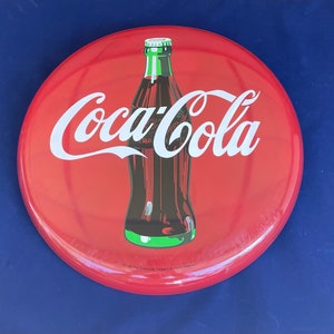 May include: Red Coca-Cola metal sign with a white Coca-Cola logo and a bottle of Coca-Cola in the center. The bottle is green and has a red cap. The sign has the text "1995 The Coca-Cola Company. All Rights Reserved."