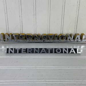 May include: Two vintage "INTERNATIONAL" emblems. The top emblem has yellow lettering with a silver border. The bottom emblem is silver with black lettering. Both are displayed against a white, wood-paneled background.