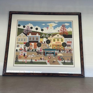 May include: A framed print of a colorful illustration depicting a small town with houses, shops, and people. The scene is set on a sunny day with a blue sky and white clouds. There are hot air balloons in the sky. The print is in a brown frame.
