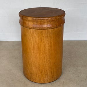 May include: A cylindrical wooden container with a dark brown lid. The body of the container is a light brown color with vertical wood grain. The lid is a darker shade of brown and has a rounded edge. The container is likely used for storage.