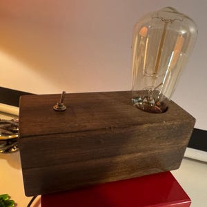 Op de afbeelding: Een handgemaakte houten lamp met een vintage Edison-stijl gloeilamp. De lamp heeft een donkerbruine rechthoekige basis en een kleine tuimelschakelaar. De heldere lamp heeft zichtbare filamenten, wat een warme gloed creëert. Een uniek woondecoratie-item.
