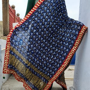 May include: A navy blue and gold patterned scarf with a red border and gold leaf design. The scarf has a geometric pattern with white accents. The edges are trimmed with gold leaf and red fabric. A green tassel hangs from the corner.