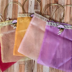 May include: Five sheer fabric tote bags in shades of red, gold, peach, pink, and purple. Each bag has a gold handle and is trimmed with decorative trim and floral accents. The bags are arranged on a wooden surface.