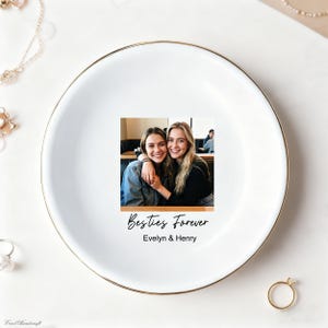 以下が含まれることがあります： 金色の縁が付いた白いセラミック製の小物入れ。皿には、「Besties Forever」の文字と「Evelyn & Henry」の名前が入った、笑顔の2人の写真が写っています。