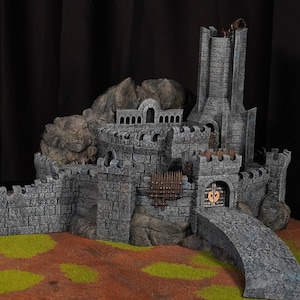 May include: A detailed model of a stone castle with a tall tower, arched gateways, and a drawbridge. The castle is gray with brown accents and sits on a base of green and brown terrain. The model is designed for tabletop gaming.