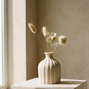 May include: A beige ceramic vase with vertical ribbed detailing holds dried flowers. The vase sits on a square, light-colored stone block. The arrangement includes fluffy, tan-colored stems and delicate white flowers.