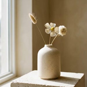 Könnte beinhalten: Eine beige gesprenkelte Keramikvase mit Trockenblumen, darunter eine weiße, gänseblümchenartige Blüte und flauschiges, hellbraunes Gras. Die Vase steht auf einem quadratischen, strukturierten Betonblock.