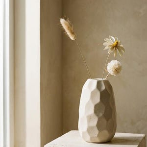 Può includere: Un vaso beige geometrico con fiori secchi. Il vaso ha un motivo a nido d'ape strutturato. I fiori includono un'erba soffice e due fiori bianchi e gialli. Il vaso è posto su un blocco di pietra, con una finestra sullo sfondo.