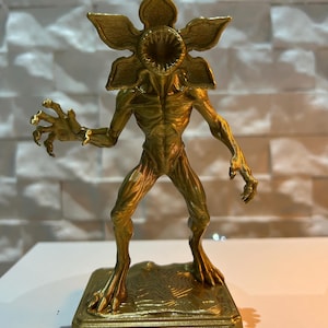 Puede incluir: Figura dorada de Demogorgon de la serie Stranger Things. La figura tiene una cabeza en forma de flor con una boca abierta, brazos extendidos y un cuerpo texturizado. La base de la figura dice "Demogorgon".