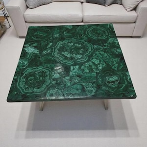 Könnte beinhalten: Quadratischer Couchtisch mit polierter Malachitplatte. Der Tisch hat eine lebendige grüne Farbe mit komplizierten Wirbelmustern. Er hat ein modernes Design mit silbernen Metallbeinen und steht auf einem weißen Teppich.