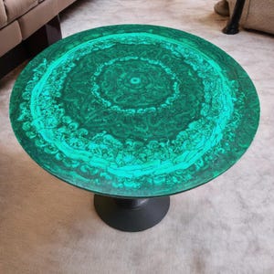 Könnte beinhalten: Runder Couchtisch mit einer lebendigen grünen Malachitplatte. Die Tischplatte weist ein wirbelndes Muster aus hellen und dunklen Grüntönen auf. Der Tisch wird von einem einzelnen, schwarzen, zylindrischen Sockel getragen.