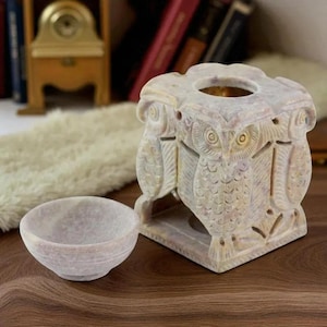 Può includere: Un portacandele in pietra scolpita a tema gufo e una piccola ciotola. Il portacandele è quadrato con intagli di gufi su ogni lato. La ciotola è rotonda e si trova accanto al portacandele. Entrambi gli oggetti sono di colore beige chiaro.