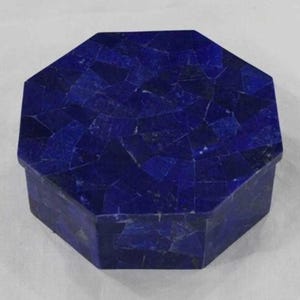 Op de afbeelding: Een donkerblauwe, achthoekige doos met een mozaïekachtig patroon. De doos is gemaakt van een diepblauw materiaal, waarbij elk stukje van het mozaïek enigszins in tint varieert. Het deksel past perfect op de basis, waardoor een decoratief opbergitem ontstaat.