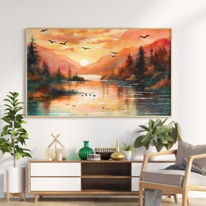 Puede incluir: Pintura de acuarela enmarcada de una puesta de sol sobre un lago, con montañas al fondo. El cielo es una mezcla de tonos naranja, amarillo y rojo. Aves silueteadas vuelan por el cielo. La pintura se exhibe sobre un mueble blanco y de madera.