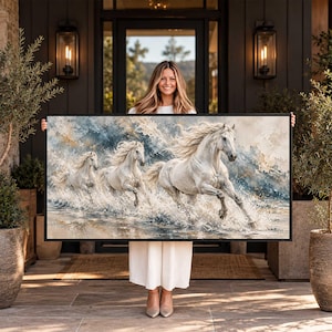 May include: A rectangular framed wall art print featuring three white horses galloping through shallow water. The horses have flowing manes and tails, depicted in a painterly style with splashes of blue, grey, and cream tones in the background.