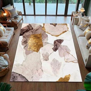 Puede incluir: Una alfombra rectangular grande con un diseño abstracto. La alfombra presenta un fondo blanco con formas superpuestas en tonos dorados, marrones y grises. La alfombra está colocada sobre un suelo de madera en un salón.