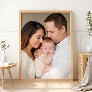 Puede incluir: Retrato familiar enmarcado con madre, padre y bebé. El bebé está acurrucado en los brazos de los padres, todos vestidos de blanco. El marco es de madera clara y el fondo es neutro.