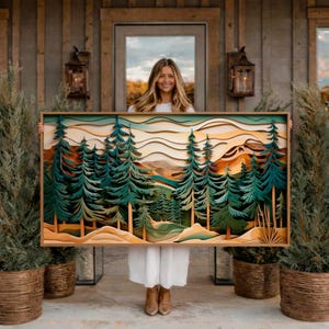 Puede incluir: Una obra de arte de madera en capas que representa una escena de bosque con altos árboles verdes, montañas y un cielo en capas. La obra de arte está enmarcada en un tono de madera natural y sostenida por una persona. La paleta de colores incluye verdes, marrones y naranjas.