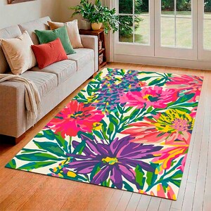 Puede incluir: Una alfombra floral rectangular con un diseño vibrante. La alfombra presenta grandes flores coloridas en tonos de rosa, morado, amarillo y azul sobre un fondo crema. Hojas y tallos verdes se intercalan en el diseño.
