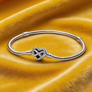 Könnte beinhalten: Ein silbernes Armband mit einem herzförmigen Anhänger mit einem Unendlichkeitssymbol. Der Anhänger ist mit kleinen, klaren Kristallen und einem schwarzen Mittelteil besetzt. Das Armband wird auf einer gelben, strukturierten Oberfläche präsentiert.
