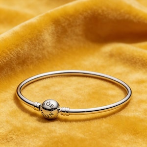 May include: A silver bracelet with a branded round charm. The bracelet is set against a yellow velvet backdrop. The design is simple and elegant, suitable for everyday wear.
