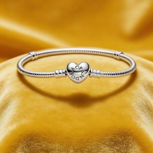 Puede incluir: Una pulsera plateada con un cierre en forma de corazón. El cierre de corazón tiene grabada la palabra "Pandora". La pulsera está sobre una superficie amarilla.