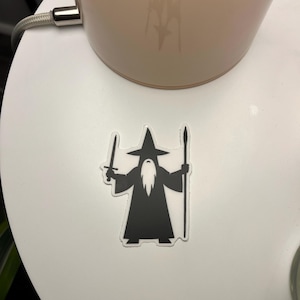 May include: A black and white sticker featuring a wizard with a long beard, holding a sword and a staff. The wizard wears a tall hat and a long robe. The sticker is on a white surface.