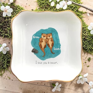 Op de afbeelding: Een vierkant wit keramieken schaaltje met een goudkleurige rand. Het midden is voorzien van een aquarelillustratie van twee bruine otters die in blauw water drijven, met de namen Chris en Emma en de tekst I love you a lotter.