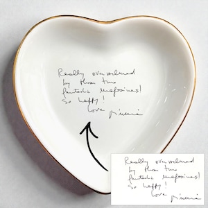 Puede incluir: Un plato de cerámica blanco en forma de corazón con un borde dorado. El plato presenta texto escrito a mano en tinta negra que dice: "Realmente abrumada por esas dos fantásticas revistas! ¡Tan feliz! Amor, Pincera."