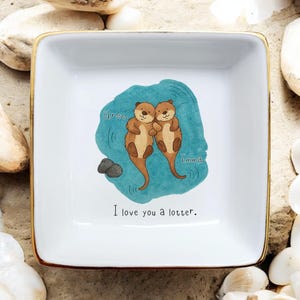 Op de afbeelding: Witte vierkante keramische schaal met een gouden rand. De schaal heeft een turquoise blauw aquarelontwerp met twee cartoonotters die elkaar omhelzen. De tekst "I love you a lotter" staat eronder.