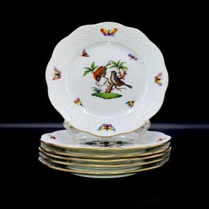 May include: A stack of white porcelain plates with scalloped edges and gold rims. The center of each plate features a colorful illustration of two birds perched on a branch, surrounded by small butterflies scattered around the textured white border.