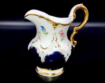 May include: A white porcelain cream jug featuring a deep cobalt blue base, ornate gold trim, and a curved gold handle. The body is decorated with small hand-painted pink and blue floral sprigs and intricate gold filigree scrollwork accents.