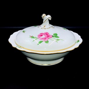 May include: A white porcelain serving bowl with a matching lid, featuring a hand-painted pink rose motif with green leaves. The bowl and lid have scalloped edges trimmed in gold, and the lid is topped with a decorative, swirling white handle.
