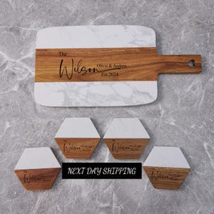 May include: A personalized serving board and matching coasters. The board features a white marble top and a wooden base with the name "The Wilson", "Olivia & Andrew", and "Est.2024". Four hexagonal coasters with the same design are included.