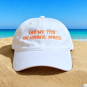 Puede incluir: Gorra de béisbol blanca con el texto "OFF MY TITS ON APEROL SPRITZ" bordado en naranja. La gorra está en una playa de arena con el mar y el cielo azul de fondo. Hay conchas marinas esparcidas alrededor de la gorra.