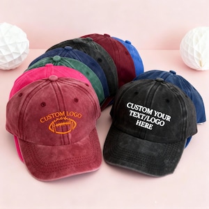 Puede incluir: Una colección de gorras de béisbol en varios colores, incluyendo negro, burdeos y azul. Algunas gorras presentan el texto "CUSTOM YOUR TEXT/LOGO HERE" en blanco, mientras que otras muestran un diseño de fútbol americano. Las gorras están dispuestas sobre una superficie rosa.