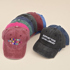 May include: A selection of baseball caps in shades like burgundy, pink, and black. The burgundy cap has the embroidered text "SUPPORT YOUR LOCAL LIBRARY" in multiple colours. The black cap shows the same message in white.