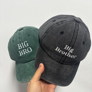 May include: Two baseball caps are shown, one dark green and one charcoal grey. The green cap has "BIG BRO" embroidered in white, while the grey cap says "Big Brother". Both caps are made of a soft, worn fabric.
