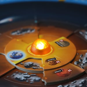 May include: A close-up of a board game depicting the solar system. The game features a central, illuminated sun and circular planet pieces labeled with planet names. The planets include Earth, Venus, Mars, and Saturn.