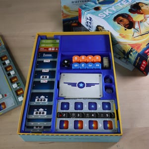 Può includere: Una scatola di gioco da tavolo aperta con componenti blu, gialli e arancioni. La scatola contiene dadi, carte e pedine. La scatola del gioco è etichettata "SKY TEAM" con immagini di persone e un aereo. È visibile un tabellone di gioco separato.