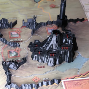 Puede incluir: Un juego de mesa con piezas de terreno de plástico negro, que incluyen un volcán y estructuras de fortaleza, colocadas sobre un mapa. El mapa presenta nombres de lugares como Morannon y Minas Morgul. Se ven cartas en el fondo.