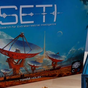 Peut inclure: Présentation du jeu de société "SETI: Search for Extraterrestrial Intelligence". L'illustration de la boîte montre des radiotélescopes sous un ciel bleu avec la lune. La boîte de jeu ouverte révèle des composants.