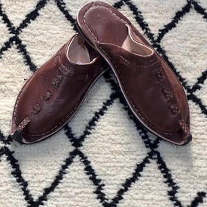 Peut inclure: Une paire de pantoufles en cuir marron avec des bouts pointus et des coutures décoratives. Les pantoufles sont présentées sur un tapis blanc avec un motif géométrique noir. Les pantoufles semblent faites à la main.